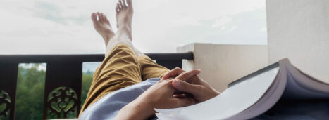 Young man reading a book lying on soft mattress in relaxing bed at terrace with green nature view. Fresh air in the morning of weekend or free day. Relax or education background idea. Selective focus.