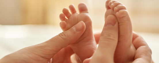 Mum making baby massage, mother massaging infant bare foot, preventive massage for newborn, mommy stroking the baby's feet with both hands on light background.