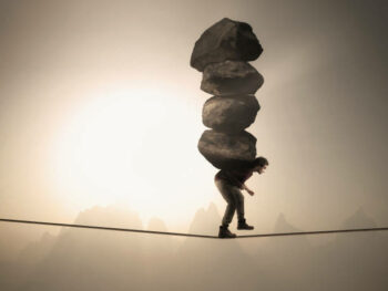 Man carries a stack of big rocks while balancing on a rope at high altitude .